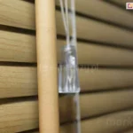 Zdjęcie przedstawia zbliżenie na detale techniczne, jakimi charakteryzują się żaluzje aluminiowe 25 mm z powłoką drewnopodobną w jasnym odcieniu dębu lub sosny. Lamele mają wyraźnie zaznaczony rysunek słojów drewna, co nadaje im naturalny wygląd przy zachowaniu trwałości metalu. W centralnej części kadru widoczne są elementy sterujące systemem osłonowym, w tym pionowe pokrętło o cylindrycznym kształcie służące do regulacji kąta nachylenia listew oraz biały sznurek zakończony transparentnym, plastikowym dzwonkiem uchwytu. Poszczególne poziome lamele o szerokości dwudziestu pięciu milimetrów są ułożone kaskadowo, lekko nachodząc na siebie, co pozwala na precyzyjne kontrolowanie ilości wpadającego światła. Konstrukcja opiera się na białej drabince sznurkowej, która przechodzi przez otwory w aluminium, utrzymując stabilność i równomierne odstępy między segmentami żaluzji. Tło po prawej stronie zdjęcia jest rozmyte, co skupia uwagę na fakturze materiału oraz mechanizmie operacyjnym produktu.