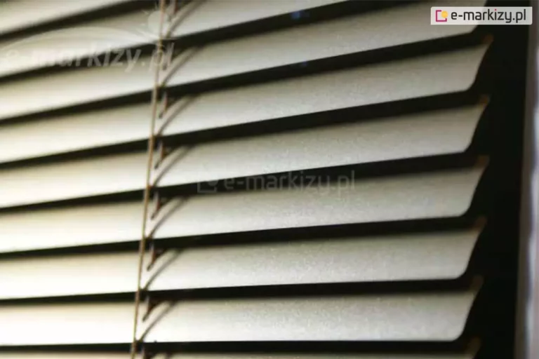 Żaluzja pozioma aluminiowa z wąskimi lamelami w kolorze srebrnym oraz drabinką sznurkową w kolorze zbliżonym do brązu zamocowana na oknie, za którym panuje ciemność.