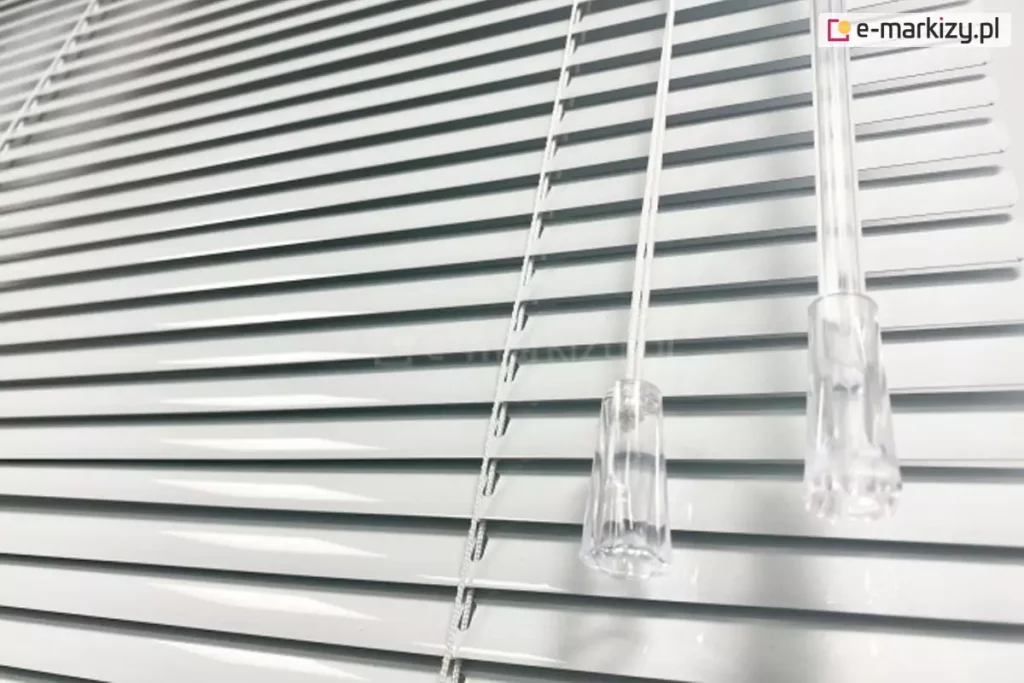 Białe żaluzje aluminiowe z lamelami 25mm o dużym połysku z białą drabinką sznurkową. Po prawej stronie widoczny sznurek do sterowania zakończony lexanowym dzwonkiem oraz pokrętło wykonana z lexanu do zmiany kąta nachylenia lameli.