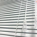 Białe żaluzje aluminiowe z lamelami 25mm o dużym połysku z białą drabinką sznurkową. Po prawej stronie widoczny sznurek do sterowania zakończony lexanowym dzwonkiem oraz pokrętło wykonana z lexanu do zmiany kąta nachylenia lameli.