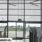 Wnętrze nowoczesnego budynku z wysoką, dwukondygnacyjną przeszkloną fasadą frontową. Na dużych, pionowych powierzchniach okiennych zamontowane są zewnętrzne żaluzje fasadowe z poziomymi lamelami w kolorze srebrnym. Lamele są równomiernie rozstawione, częściowo uchylone, co pozwala na regulację dopływu światła dziennego do wnętrza pomieszczenia, jednocześnie zapewniając widoczność na zewnątrz. W centralnej części kadru znajduje się wyjście ewakuacyjne – przeszklone, dwuskrzydłowe drzwi z zielonym oznaczeniem „wyjście ewakuacyjne” powyżej. Po lewej stronie widoczna jest część stanowiska recepcyjnego z osobą siedzącą przy biurku. Po prawej stronie umieszczone są metalowe stojaki ekspozycyjne z tekstyliami lub odzieżą roboczą. W tle przez szyby widać otwartą przestrzeń z kostką brukową oraz zabudową mieszkalną i przemysłową. Na suficie zamontowano nowoczesne, poziome oprawy oświetleniowe w układzie liniowym.