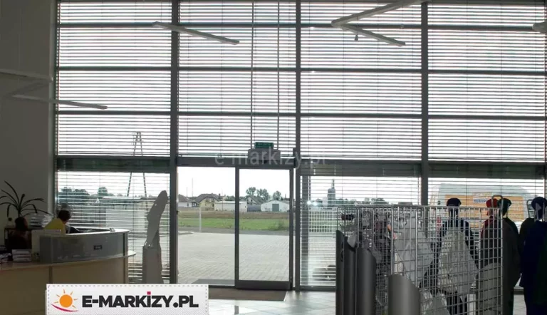Wnętrze nowoczesnego budynku z wysoką, dwukondygnacyjną przeszkloną fasadą frontową. Na dużych, pionowych powierzchniach okiennych zamontowane są zewnętrzne żaluzje fasadowe z poziomymi lamelami w kolorze srebrnym. Lamele są równomiernie rozstawione, częściowo uchylone, co pozwala na regulację dopływu światła dziennego do wnętrza pomieszczenia, jednocześnie zapewniając widoczność na zewnątrz. W centralnej części kadru znajduje się wyjście ewakuacyjne – przeszklone, dwuskrzydłowe drzwi z zielonym oznaczeniem „wyjście ewakuacyjne” powyżej. Po lewej stronie widoczna jest część stanowiska recepcyjnego z osobą siedzącą przy biurku. Po prawej stronie umieszczone są metalowe stojaki ekspozycyjne z tekstyliami lub odzieżą roboczą. W tle przez szyby widać otwartą przestrzeń z kostką brukową oraz zabudową mieszkalną i przemysłową. Na suficie zamontowano nowoczesne, poziome oprawy oświetleniowe w układzie liniowym.