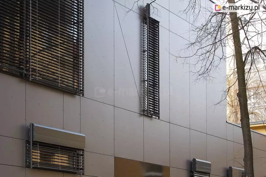 Systemy osłon okiennych zamontowane na technicznej elewacji budynku wykonanej z ciemnoszarych paneli kompozytowych z widocznymi nitami montażowymi. Zdjęcie prezentuje różne typy i rozmiary żaluzji fasadowych: od dużych powierzchni przesłaniających okna po wąskie, pionowe pasy żaluzji oraz mniejsze moduły z widocznymi kasetami osłonowymi typu Oval, montowanymi natynkowo, z prowadnicami umieszczonymi po bokach kaset. Ciemne lamele kontrastują z matową strukturą fasady. Po prawej stronie widoczne są gałęzie drzewa pozbawionego liści, co sugeruje okres jesienno-zimowy i pokazuje integrację systemów osłonowych z otoczeniem miejskim.