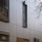 Systemy osłon okiennych zamontowane na technicznej elewacji budynku wykonanej z ciemnoszarych paneli kompozytowych z widocznymi nitami montażowymi. Zdjęcie prezentuje różne typy i rozmiary żaluzji fasadowych: od dużych powierzchni przesłaniających okna po wąskie, pionowe pasy żaluzji oraz mniejsze moduły z widocznymi kasetami osłonowymi typu Oval, montowanymi natynkowo, z prowadnicami umieszczonymi po bokach kaset. Ciemne lamele kontrastują z matową strukturą fasady. Po prawej stronie widoczne są gałęzie drzewa pozbawionego liści, co sugeruje okres jesienno-zimowy i pokazuje integrację systemów osłonowych z otoczeniem miejskim.
