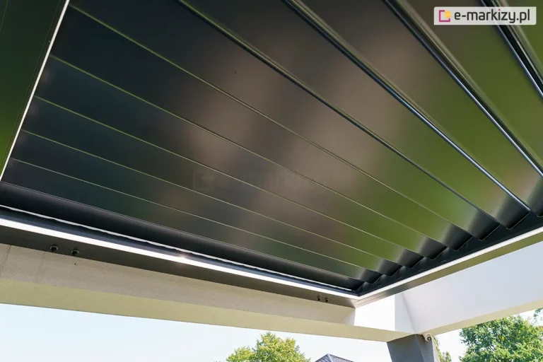 widok od dołu na w pełni zamknięty dach lamelowy pergoli SB400R w kolorze antracytowym. Konstrukcja zadaszenia składa się z szeregu płaskich, aluminiowych piór (lameli), które po obróceniu tworzą jednolitą i szczelną płaszczyznę chroniącą przed opadami oraz słońcem. Powierzchnia lameli charakteryzuje się matowym wykończeniem z delikatnym satynowym połyskiem, w którym odbija się naturalne światło. Zadaszenie jest osadzone w solidnej ramie konstrukcyjnej, która od wewnętrznej strony została wyposażona w zintegrowane oświetlenie liniowe LED, widoczne jako jasny pas światła wzdłuż krawędzi. Całość zamontowana jest do jasnej elewacji budynku i wsparta na betonowym lub tynkowanym słupie w kolorze białym. W dolnej części kadru widoczny jest fragment błękitnego nieba oraz korony zielonych drzew, co podkreśla nowoczesną estetykę systemu wkomponowanego w otoczenie ogrodu.