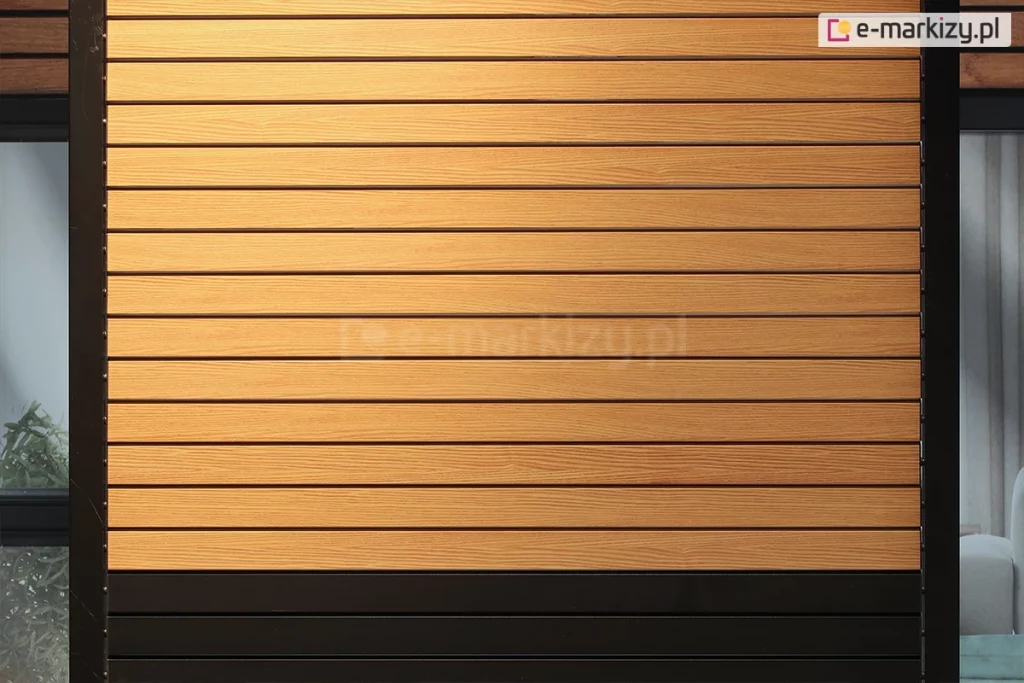 Poziome lamele wykonane z aluminium w okleinie drewnopodobnej o wyraźnym usłojeniu w kolorze jasnego dębu stanowiące wypełnienie nowoczesnego systemu osłonowego. Każdy panel posiada subtelne żłobienia imitujące naturalną strukturę drewna, a ich regularne rozmieszczenie tworzy rytmiczną, uporządkowaną płaszczyznę. Konstrukcja osadzona jest w masywnej, pionowej ramie bocznej o matowym wykończeniu w kolorze czarnym lub bardzo ciemnym antracycie. W dolnej części przesłony widoczny jest fragment segmentu w jednolitym, czarnym kolorze, który kontrastuje z ciepłym odcieniem górnych paneli. Po bokach ramy dostrzec można elementy montażowe w formie prowadnic lub linek bocznych. W tle za przesłoną widoczne są fragmenty przeszkleń okiennych, zarys mebli ogrodowych oraz zielona roślinność, co sugeruje zastosowanie produktu jako bocznej ściany pergoli, tarasu lub altany zapewniającej prywatność i ochronę przeciwsłoneczną. Całość charakteryzuje się nowoczesnym designem łączącym estetykę naturalnych materiałów z trwałością systemów metalowych.