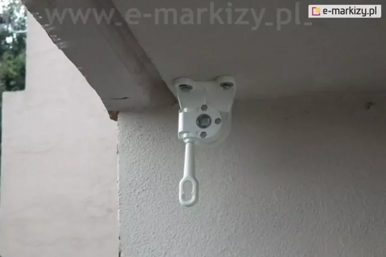 Zdjęcie przedstawia biały, aluminiowy uchwyt montażowy markizy balkonowej-poręczowej zamocowany do sufitu wnęki balkonowej, wyposażony w przekładnię oczkową do obsługi ręcznej. Wspornik jest solidnie przytwierdzony do jasnej, otynkowanej powierzchni za pomocą śrub montażowych widocznych u nasady. Kluczowym elementem widocznym na tym ujęciu jest mechanizm przekładni w kolorze białym, zamontowany bezpośrednio w gnieździe uchwytu. Z mechanizmu wyprowadzone jest pionowo w dół białe "oczko" (pętla), które służy do zaczepienia korby manualnej, umożliwiającej rozwijanie i zwijanie markizy. Sama obudowa przekładni posiada widoczne otwory technologiczne oraz śruby mocujące mechanizm do ramienia wspornika. Konstrukcja znajduje się blisko narożnika ściany o porowatej strukturze tynku. W tle widoczny jest fragment jasnej elewacji budynku oraz rozmyte zielone liście drzew, co sugeruje ekspozycję zewnętrzną na wysokości piętra. Zdjęcie wykonane jest pod kątem, podkreślając funkcjonalny aspekt mechanizmu sterującego markizą pionową lub balkonową.