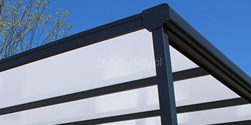 Zdjęcie prezentuje zbliżenie na narożnik aluminiowego zadaszenia tarasowego Nebbiolo Deponti w kolorze ciemnoantracytowym. Głównym elementem technicznym jest estetyczna zaślepka rynny zamontowana na zakończeniu profilu czołowego, precyzyjnie dopasowana do kształtu ramy i przykręcona za pomocą widocznych śrub. Konstrukcja wspiera zadaszenie wykonane z mlecznych płyt poliwęglanowych, które są osadzone w równoległych profilach krokwiowych. Ujęcie ukazuje wysoką jakość wykończenia detali oraz szczelność systemu odprowadzania wody deszczowej. W tle widoczny jest fragment korony drzewa z młodymi liśćmi oraz czyste, błękitne niebo, co podkreśla kontrast między techniczną strukturą aluminium a naturalnym otoczeniem.