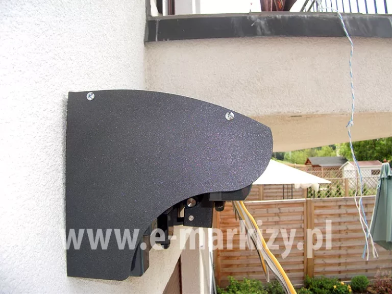Uniwersalny daszek do markiz w kolorze srebrzysty grafit z widocznymi śrubami mocującymi blachę górną zawieszony na ścianie z białym tynkiem strukturalnym w pobliżu balkonu, z którego zwisa biało-niebieski sznurek, w tle widoczny płot oraz parasol przeciwsłoneczny i pawilon z białym dachem