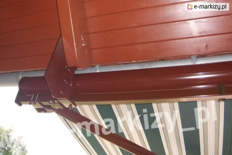Detaliczne zdjęcie prezentujące techniczne rozwiązanie montażowe markizy tarasowej Palladio marki Aluprof z wykorzystaniem specjalnego uchwytu krokwiowego. Metalowy uchwyt w kształcie odwróconej litery L został przykręcony bezpośrednio do boku drewnianej krokwi dachowej, która wraz z widocznym powyżej podbitkiem została pomalowana lakierobejcą w kolorze ciepłego brązu. Elementy montażowe, w tym korpus uchwytu, profilowana kaseta markizy oraz ramiona przegubowe, wykończono lakierem proszkowym w dopasowanym, czekoladowym odcieniu brązu. Na pionowej ściance uchwytu krokwiowego oraz na dolnych mocowaniach konstrukcji widoczne są precyzyjnie założone, okrągłe zaślepki śrub, które estetycznie maskują łby łączników mechanicznych i zabezpieczają je przed korozją. Pod kasetą ochronną znajduje się częściowo rozwinięte poszycie markizy wykonane z tkaniny w podłużne pasy, gdzie dominują szerokie pasy w kolorze ciemnej zieleni butelkowej oraz węższe, symetryczne paski w odcieniach beżu, szarości i bieli. W tle, tuż za kasetą, przebiega poziomo szara rynna dachowa, a w lewym dolnym rogu kadru widać zarys zielonych liści drzew na zewnątrz. Zdjęcie wykonano z dolnej perspektywy, skupiając uwagę na solidności instalacji oraz detalach wykończenia maskującego.