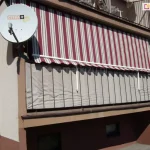 Zdjęcie ukazuje elewację bloku mieszkalnego z wyraźnie wyeksponowanym balkonem na parterze, na którym zamontowana jest markiza balkonowa poręczowa. Poszycie markizy charakteryzuje się wzorem w pionowe, naprzemienne pasy w kolorach bordowym, kremowym oraz wąskimi szarymi akcentami. Tkanina jest rozwinięta i zwisa pionowo w dół, opierając się na białych, metalowych hakach w ramionach przytwierdzonych do barierki. Dolna krawędź materiału zakończona jest falbaną o falistym kształcie z białą lamówką. Balustrada balkonu, wykonana z ciemnych profili, została przesłonięta beżową matą osłonową, która szczelnie wypełnia przestrzeń między szczeblami. Po lewej stronie balkonu, na bocznej ścianie budynku, znajduje się okrągła, biała antena satelitarna z logo operatora. Elewacja budynku jest dwukolorowa: górna część ma jasny, beżowy odcień, natomiast dolna partia pod balkonem oraz fundamenty są pomalowane na kolor ciemnobrązowy. W tle widać dalszą część bloku z rzędem okien i balkonów oraz fragment zielonego trawnika. Światło słoneczne pada z prawej strony, rzucając wyraźny cień markizy na wnękę balkonową i matę osłonową.