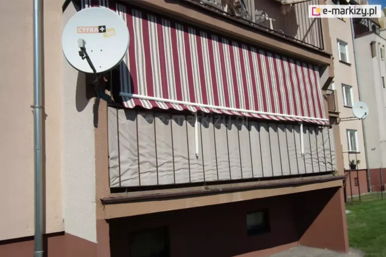 Zdjęcie ukazuje elewację bloku mieszkalnego z wyraźnie wyeksponowanym balkonem na parterze, na którym zamontowana jest markiza balkonowa poręczowa. Poszycie markizy charakteryzuje się wzorem w pionowe, naprzemienne pasy w kolorach bordowym, kremowym oraz wąskimi szarymi akcentami. Tkanina jest rozwinięta i zwisa pionowo w dół, opierając się na białych, metalowych hakach w ramionach przytwierdzonych do barierki. Dolna krawędź materiału zakończona jest falbaną o falistym kształcie z białą lamówką. Balustrada balkonu, wykonana z ciemnych profili, została przesłonięta beżową matą osłonową, która szczelnie wypełnia przestrzeń między szczeblami. Po lewej stronie balkonu, na bocznej ścianie budynku, znajduje się okrągła, biała antena satelitarna z logo operatora. Elewacja budynku jest dwukolorowa: górna część ma jasny, beżowy odcień, natomiast dolna partia pod balkonem oraz fundamenty są pomalowane na kolor ciemnobrązowy. W tle widać dalszą część bloku z rzędem okien i balkonów oraz fragment zielonego trawnika. Światło słoneczne pada z prawej strony, rzucając wyraźny cień markizy na wnękę balkonową i matę osłonową.