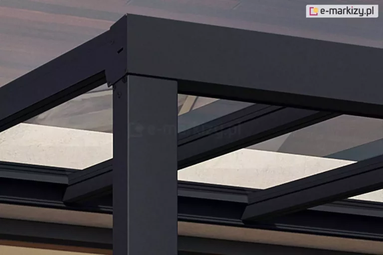 Zdjęcie prezentuje techniczne zbliżenie na górny narożnik konstrukcji zadaszenia tarasowego Modes Glas w kolorze antracytowym. Widoczne jest precyzyjne połączenie pionowego słupa nośnego z poziomą belką rynnową oraz prostopadle biegnącą belką boczną. Zewnętrzny styk elementów został estetycznie zabezpieczony dopasowaną, płaską maskownicą przykręconą dyskretnymi wkrętami. Wewnątrz konstrukcji widoczne są systemowe profile krokwiowe z czarnymi uszczelkami, w których osadzono panele dachowe ze szkła bezpiecznego.