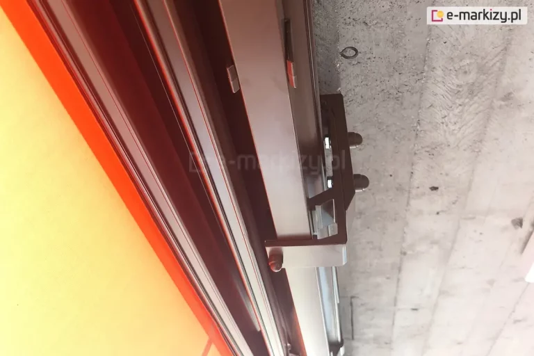 Zdjęcie przedstawia szczegółowy widok techniczny na system mocowania markizy, prezentujący zespolenie uchwytu ściennego i sufitowego marki Aluprof. Na pierwszym planie widoczne są solidne, metalowe profile konstrukcyjne w kolorze ciemnobrązowym, które są precyzyjnie połączone za pomocą śrub zabezpieczonych estetycznymi kapturkami ochronnymi. Elementy montażowe są przytwierdzone bezpośrednio do surowej, betonowej powierzchni stropu o wyraźnej teksturze. Po lewej stronie kadru znajduje się fragment napiętej tkaniny poszycia w kolorze intensywnie pomarańczowym lub żółtym, która jest osadzona w prowadnicy mechanizmu. Fotografia wykonana z bliskiej perspektywy ukazuje stabilność i masywność konstrukcji wsporczej, umożliwiającej bezpieczny montaż markizy w trudnych warunkach przestrzennych.