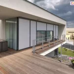 Przestronny taras przynależący do nowoczesnego apartamentowca, wykończony deskami kompozytowymi w ciepłym, naturalnym odcieniu drewna. Całość utrzymana jest w minimalistycznej estetyce z naciskiem na proste linie, kontrasty kolorystyczne i funkcjonalność. Na elewacji zamontowane zostały eleganckie refleksole z serii Refleksol 103 marki Selt, w jasnoszarym odcieniu, doskonale dopasowanym do antracytowych ram okiennych i ciemnych profili konstrukcyjnych. Rolety pokrywają całą powierzchnię dużych przeszkleń, nadając elewacji jednolity, nowoczesny wygląd. W lewym rogu tarasu widoczny jest kompaktowy zestaw mebli ogrodowych z technorattanu – czarny stolik ze szklanym blatem i wsunięte pod niego dwa fotele. Po prawej stronie, przy barierce, stoi donica z kolorowymi pelargoniami, które ożywiają neutralną kolorystykę przestrzeni i dodają jej domowego charakteru. Balustrada z metalową ramą zapewnia niezakłócony widok na zieloną przestrzeń ogrodu i sąsiednie budynki. W głębi widać ukrytą strefę wypoczynkową z narożnikiem i piecem, a na horyzoncie niebo pokryte jest dynamicznymi chmurami, co wprowadza nieco dramatyzmu i kontrastuje z uporządkowaną, spokojną kompozycją tarasu.