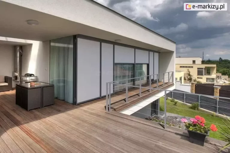 Przestronny taras przynależący do nowoczesnego apartamentowca, wykończony deskami kompozytowymi w ciepłym, naturalnym odcieniu drewna. Całość utrzymana jest w minimalistycznej estetyce z naciskiem na proste linie, kontrasty kolorystyczne i funkcjonalność. Na elewacji zamontowane zostały eleganckie refleksole z serii Refleksol 103 marki Selt, w jasnoszarym odcieniu, doskonale dopasowanym do antracytowych ram okiennych i ciemnych profili konstrukcyjnych. Rolety pokrywają całą powierzchnię dużych przeszkleń, nadając elewacji jednolity, nowoczesny wygląd. W lewym rogu tarasu widoczny jest kompaktowy zestaw mebli ogrodowych z technorattanu – czarny stolik ze szklanym blatem i wsunięte pod niego dwa fotele. Po prawej stronie, przy barierce, stoi donica z kolorowymi pelargoniami, które ożywiają neutralną kolorystykę przestrzeni i dodają jej domowego charakteru. Balustrada z metalową ramą zapewnia niezakłócony widok na zieloną przestrzeń ogrodu i sąsiednie budynki. W głębi widać ukrytą strefę wypoczynkową z narożnikiem i piecem, a na horyzoncie niebo pokryte jest dynamicznymi chmurami, co wprowadza nieco dramatyzmu i kontrastuje z uporządkowaną, spokojną kompozycją tarasu.