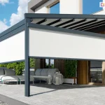 Aluminiowa pergola tarasowa w kolorze ciemnego antracytu, zamontowana do elewacji nowoczesnego budynku mieszkalnego, charakteryzującego się połączeniem bieli oraz pionowych paneli o strukturze jasnego drewna. Konstrukcja zadaszenia opiera się na trzech solidnych, pionowych słupach o kwadratowym przekroju, ustawionych na jasnoszarym, betonowym podłożu tarasu. Górna część pergoli posiada ruchome poszycie tkaninowe w formie jasnych pasów, które są rozpięte pomiędzy belkami przeciwwietrznymi. Frontowa oraz boczna strona pergoli zostały wyposażone w pionowe osłony typu screen (refleksol Zip L Zip U), z tkaniną screen w kolorze białym, które są częściowo rozwinięte, zasłaniając górną połowę przestrzeni między słupami i tworząc barierę przed słońcem. Pod zadaszeniem znajduje się przestronna strefa wypoczynkowa z nowoczesnymi meblami ogrodowymi, w tym niskimi sofami z grubymi, jasnoszarymi poduchami oraz białymi ażurowymi lampionami dekoracyjnymi. W tle widoczne są duże przeszklenia okienne, w których odbija się błękitne niebo z białymi chmurami. Po lewej stronie kadru znajduje się fragment basenu z błękitną wodą, oddzielony od tarasu pasem drobnego, szarego żwiru. Całość kompozycji dopełnia niska roślinność w donicach oraz soczysta zieleń drzew widoczna w tle posesji.
