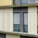 Nowoczesna elewacja budynku biurowego lub mieszkalnego wykonana z pionowych paneli w kolorze piaskowym oraz ciemnoszarym. Na fasadzie widoczne są cztery okna wyposażone w zewnętrzne refleksole 120 Aluprof. z tkaniną screen w kolorze szarym. Rolety są częściowo opuszczone do około 3/4 wysokości okna, odsłaniając dolne przeszklenia, w pasy matowego i przejrzystego szkła. Tkanina rolet charakteryzuje się drobnym splotem zapewniającym ochronę przeciwsłoneczną przy zachowaniu częściowej przezierności. Systemy osłonowe są zintegrowane z elewacją, z widocznymi prowadnicami bocznymi w kolorze ciemnoszarym, dopasowanymi do stolarki okiennej i paneli fasadowych. Całość kompozycji podkreśla rytm i geometryczny podział nowoczesnej architektury.