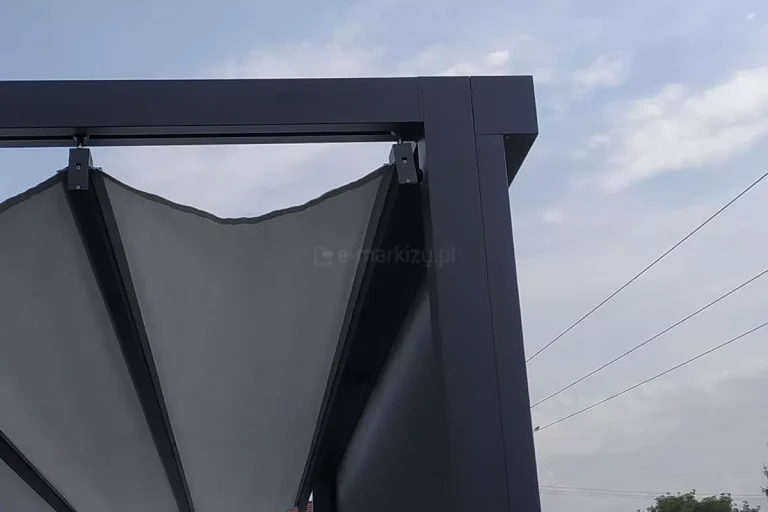Front pergoli Dragon osłonięty idealnie zlicowanym z konstrukcją pergoli refleksolem. Rozwinięty dach tkaninowy pergoli zatrzymuje się tuż przed podporami, a woda z niego spływa do boku przez widoczne fale. W tle widoczne niebo oraz przewody wysokiego napięcia.