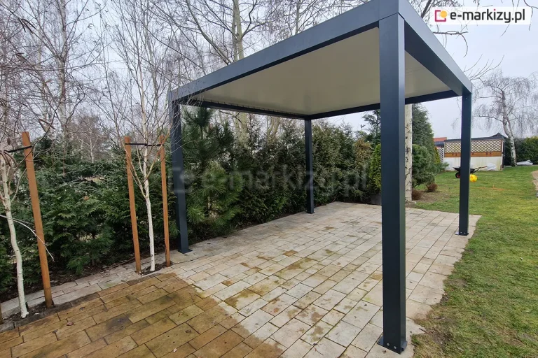 Wolnostojąca pergola lamelowa SB350 Aluprof w kolorze antracytowym, ujęta w perspektywie bocznej na terenie ogrodu. Konstrukcja z czterema pionowymi słupami nośnymi podtrzymuje płaski dach z białymi lamelami, które są całkowicie zamknięte, tworząc szczelną płaszczyznę, która skutecznie chroni przed słońcem i deszczem. Pergola stoi na podłożu z jasnej, prostokątnej kostki brukowej. Po lewej stronie widoczne są młode drzewka zabezpieczone drewnianymi palikami, a w tle gęsty żywopłot z tui, wysokie sosny oraz brzoza. W oddali po prawej stronie widać fragment trawnika z taczką oraz drewnianą architekturę ogrodową pod jasnym niebem.