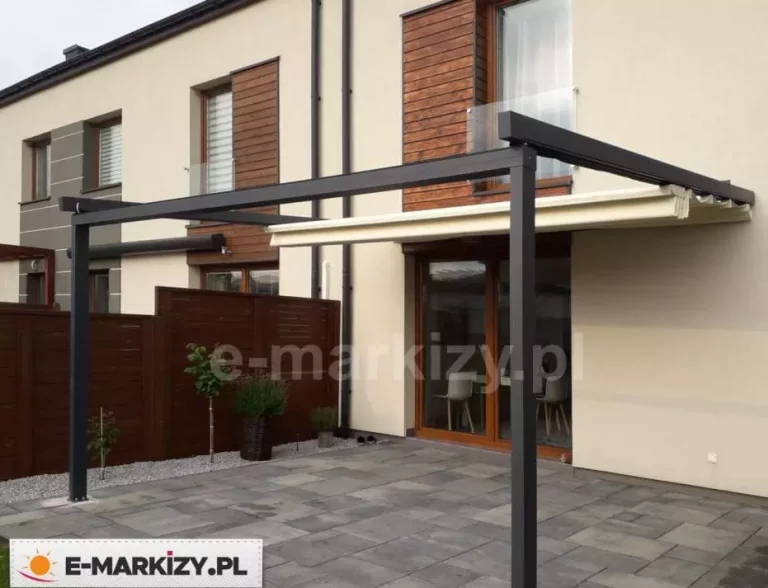 Zdjęcie przedstawia nowoczesną pergolę tarasową o konstrukcji aluminiowej w kolorze ciemnego antracytu, zamontowaną do elewacji budynku szeregowego. Zadaszenie wyposażone jest w ruchomy, zwijany dach wykonany z jasnej, kremowej tkaniny technicznej, która na zdjęciu jest częściowo zwinięta, ukazując mechanizm składania materiału w charakterystyczne fałdy. Konstrukcja opiera się na dwóch solidnych, przednich słupach o kwadratowym przekroju, przytwierdzonych do podłoża wyłożonego dużymi, szarymi płytami tarasowymi. Elewacja budynku jest jasna, z dekoracyjnymi wstawkami z drewna o pionowym usłojeniu wokół wysokich okien i drzwi tarasowych. W tle widoczne jest drewniane ogrodzenie panelowe w kolorze brązowym oraz minimalistyczne nasadzenia roślinne w donicach na pasie drobnego żwiru. Całość prezentuje estetyczne i praktyczne rozwiązanie ochrony przeciwsłonecznej w nowoczesnej zabudowie mieszkalnej.