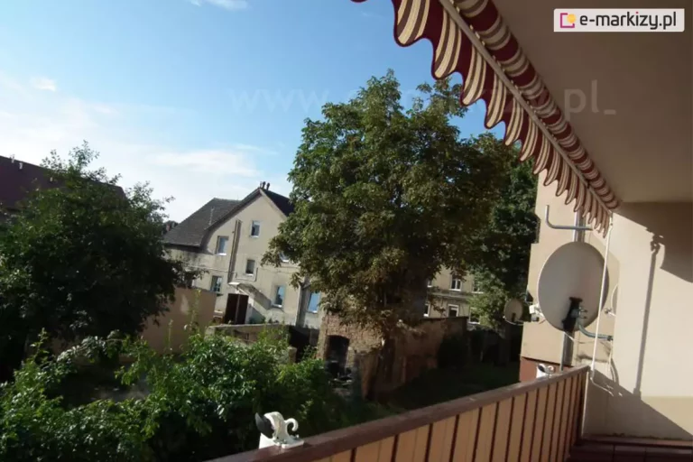Widok z balkonu na osiedle mieszkaniowe w słoneczny dzień, przedstawiający zamontowaną markizę balkonową oraz okoliczną architekturę i roślinność. W górnej części kadru widoczny jest fragment rozwiniętego poszycia markizy w szerokie, pionowe pasy w kolorach bordowym, beżowym i białym, zakończonego ozdobną falbaną o falistym brzegu z lamówką. Markiza rzuca cień na ścianę budynku po prawej stronie, na której zamontowana jest biała antena satelitarna. Balkon posiada brązową barierkę wykonaną z pionowych szczebli, a na jej poręczy widoczny jest biały element montażowy. W tle, za gęstymi zielonymi koronami drzew, znajduje się wielopiętrowy budynek mieszkalny o jasnej elewacji z licznymi oknami i dwuspadowym dachem pokrytym ciemną dachówką. Po lewej stronie widoczne są niższe zabudowania oraz fragmenty ogrodów z intensywną zielenią krzewów. Niebo nad budynkami jest bezchmurne, o jasnoniebieskim odcieniu, co sugeruje ekspozycję południową lub zachodnią i dużą intensywność światła słonecznego, przed którym chroni zamontowane zadaszenie.
