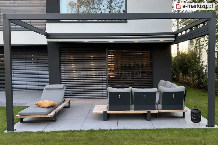 Nowoczesny taras ogrodowy w ujęciu od frontu. Taras osłania prostokątna, aluminiowa pergola w kolorze antracytowym, ze zwiniętym dachem tkaninowym, w którym zamontowano niewielkie lampki LED – włączone, rozświetlają przestrzeń pod dachem. Pod pergolą, centralnie na zdjęciu, umieszczono dwa główne meble wypoczynkowe: po lewej leżanka ogrodowa z zagłówkiem, tapicerowana jasnoszarą tkaniną i wyposażona w żółtą poduszkę dekoracyjną; po prawej narożnik z tego samego zestawu, również z jasnoszarymi poduszkami i drewnianą podstawą, z widocznymi czarnymi oparciami. Pomiędzy nimi – pusty obszar tarasu z płytkami w odcieniu szarości. W tle dom z nowoczesną elewacją i dużymi przeszkleniami, osłoniętymi poziomymi żaluzjami zewnętrznymi. Na trawie, po prawej stronie, stoi niska ozdobna latarenka z ażurową strukturą. Po bokach kadru – widoczna zieleń ogrodu i roślinność otaczająca taras. Kompozycja jest uporządkowana i utrzymana w spójnej, nowoczesnej estetyce.