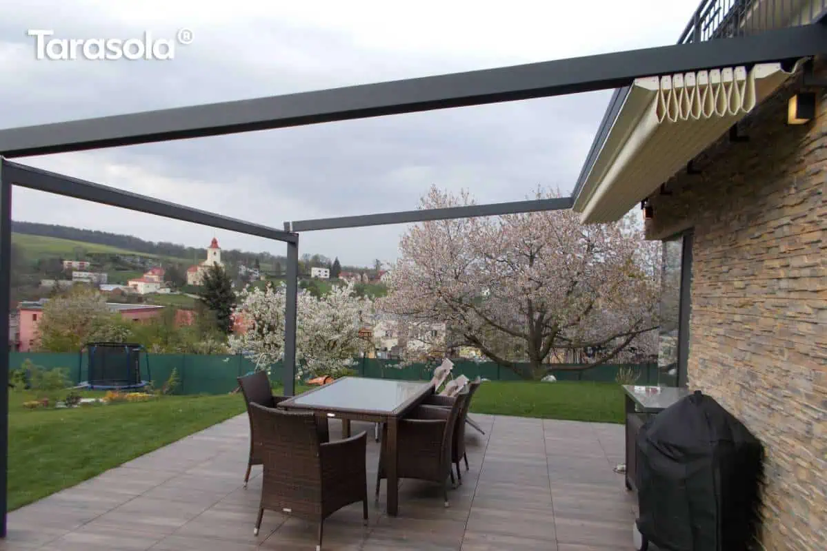 Przyścienna pergola tarasowa Elegancy Tarasola z całkowicie zwiniętym dachem rzymskim, zamontowana na tarasie wyłożonym płytkami imitującymi drewno. Antracytowa konstrukcja aluminiowa składa się z poziomych belek obwodowych oraz pionowych podpór, tworząc otwartą przestrzeń nad zestawem mebli ogrodowych. Biała tkanina zadaszenia jest złożona w ciasne pakiety pod daszkiem systemowym przy elewacji budynku wykończonej dekoracyjnym kamieniem. Na pierwszym planie widoczny jest stół z szklanym blatem otoczony technorattanowymi fotelami oraz grill pod czarnym pokrowcem. W tle rozpościera się widok na zielony ogród z kwitnącymi drzewami owocowymi, a dalej na pagórkowaty krajobraz z zabudową wiejską i kościołem pod pochmurnym niebem.