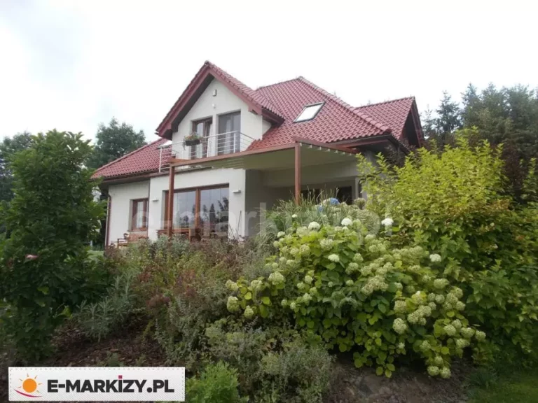 Zdjęcie przedstawia szerokie ujęcie domu jednorodzinnego z zamontowaną przyścienną pergolą tarasową Luxo. Konstrukcja zadaszenia wykonana jest z aluminium w kolorystyce drewna w brązowym odcieniu i została zamocowana bezpośrednio do jasnej elewacji budynku, wspierając się na zewnętrznych słupach nośnych. Pergola posiada ruchome, jasne poszycie tkaninowe, które w tej konfiguracji osłania znaczną część tarasu na parterze. Budynek charakteryzuje się białymi ścianami, dużymi przeszkleniami z brązowymi ramami oraz spadzistym dachem pokrytym czerwoną dachówką ceramiczną z widocznym oknem połaciowym. Na piętrze znajduje się balkon z metalową barierką, usytuowany bezpośrednio nad częścią zadaszenia. Przed tarasem rozciąga się gęsty ogród z licznymi nasadzeniami, w tym kwitnącymi na biało krzewami hortensji, ozdobnymi trawami oraz wyższymi drzewami w tle. Całość kompozycji ukazuje harmonijne połączenie nowoczesnego zadaszenia z klasyczną bryłą domu i naturalnym otoczeniem ogrodowym.
