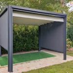 Wolnostojąca pergola tarasowa Tako o antracytowej konstrukcji aluminiowej, wyposażona w jasny, segmentowy dach tkaninowy oraz dwie rozwinięte boczne rolety typu refleksol w kolorze ciemnoszarym. Pergola jest ustawiona w ogrodzie na podłożu z jasnej kostki brukowej, na której położono pas sztucznej trawy w kolorze intensywnej zieleni. Zadaszenie jest w całości rozłożone, tworząc falowaną powierzchnię. Konstrukcja wsparta na czterech narożnych słupach znajduje się na tle gęstych, zielonych krzewów iglastych oraz drzew liściastych z żółknącymi liśćmi. Po prawej stronie widoczne są fragmenty drewnianych belek leżących na trawniku oraz kwiaty ogrodowe.