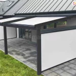 Pergola tarasowa Square ustawiona przy ścianie domku o jasnej betonowej fasadzie, nad tarasem z płyt betonowych, wyposażona w boczne osłony refleksol Zip L Zip U, które skutecznie chronią przestrzeń tarasu przed słońcem, deszczem, wiatrem i spojrzeniami sąsiadów. Przy tarasie zadbany trawnik.