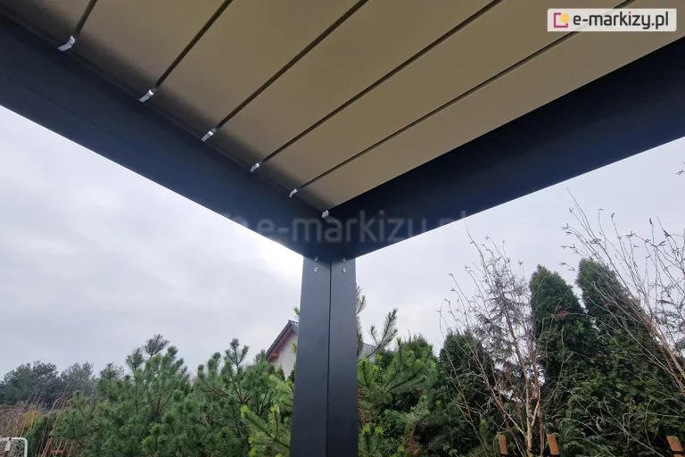 Perspektywiczne ujęcie od dołu przedstawiające narożnik pergoli SB350 Selt. Zdjęcie ukazuje połączenie pionowego słupa nośnego z poziomą ramą konstrukcyjną w kolorze antracytowym. Na słupie widoczne są cztery srebrne śruby montażowe z łbem imbusowym. Powyżej ramy znajdują się szczelnie zamknięte, białe lamele dachowe, zamocowane za pomocą metalowych uchwytów. W tle widoczna jest gęsta roślinność ogrodowa z wysokimi tujami i sosnami oraz fragment dachu sąsiedniego budynku pod zachmurzonym, jasnym niebem.