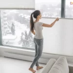 Kobieta zasłaniająca białe żaluzje do zasłonięcia dużego okna na prowadnicach sznurkowych o lekkim stopniu przejrzystości dla przestrzennego pomieszczenia