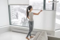 Kobieta zasłaniająca białe żaluzje do zasłonięcia dużego okna na prowadnicach sznurkowych o lekkim stopniu przejrzystości dla przestrzennego pomieszczenia