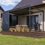 Pergola wyposażona w system częściowo otwartej rolety dachowej Veranda Selt oraz obudowana ruchomymi deseczkami aluminiowymi na tarasie z wypoczynkiem tarasowym oraz grillem