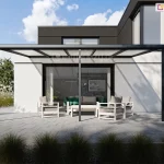 Wsparte na trzech nogach aluminiowe nowoczesne zadaszenie tarasu Nebbiolo Deponti zamontowane do czarno-białego domu osłaniające poliwęglanowym dachem drewniane meble z poduszkami