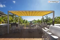 Aluminiowa pergola przeciwsłoneczna Tako z żółtym, zwijanym dachem. Wolnostojąca konstrukcja na drewnianym tarasie restauracyjnym z widokiem na tropikalny krajobraz i ocean. Eleganckie, białe meble barowe i stoliki zapewniają komfortową przestrzeń do wypoczynku