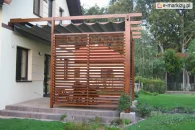 Drewniana pergola przyścienna nad wejściem do domu, wyposażona w dach rzymski Supro i ruchome deseczki shutters. Pergola osłania taras, zapewniając prywatność i ochronę przed słońcem. Drewniana konstrukcja harmonizuje z elewacją budynku oraz zielonym otoczeniem ogrodu