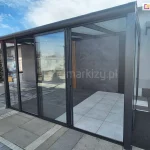 ściany przesuwne ramowe Sotro – Galeria - Całoroczny, nowoczesny ogród zimowy wyposażony w przesuwne drzwi szklane Sotro, osadzone w eleganckiej, grafitowej ramie aluminiowej. Zdjęcie ukazuje jasne, przestronne wnętrze z podłogą wyłożoną białymi i antracytowymi płytami tarasowymi.