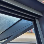 Łączenie górnej prowadnicy systemu drzwi szklanych Sotro z bocznym profilem, zamocowanym do narożnej nogi konstrukcji pergoli. Solidne aluminiowe elementy w ciemnoszarym kolorze na tle przeszklonego dachu pokrytego kroplami wody