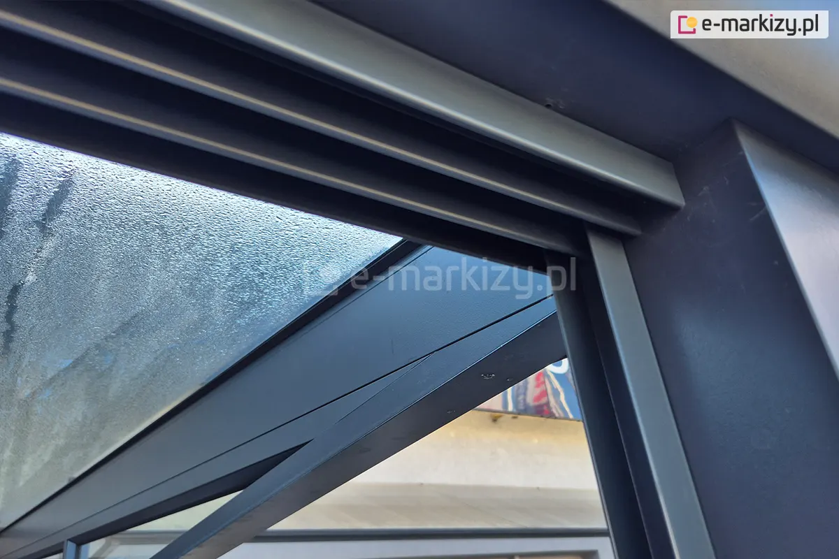 Łączenie górnej prowadnicy systemu drzwi szklanych Sotro z bocznym profilem, zamocowanym do narożnej nogi konstrukcji pergoli. Solidne aluminiowe elementy w ciemnoszarym kolorze na tle przeszklonego dachu pokrytego kroplami wody