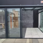 Przesuwne panele szklane Sotro z ramą aluminiową, otwarte i przesunięte na lewą stronę, prezentujące wygodny sposób dostępu do pergoli. W tle widoczne nowoczesne wnętrze pergoli o neutralnych barwach – odcieniach szarości i beżu, które tworzą elegancki, harmonijny nastrój.