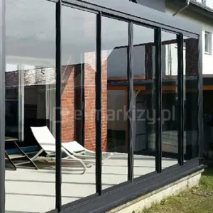 System ramowego szkła przesuwnego Sotro w kolorze czarnym zamontowany na pergoli tarasowej przy domu z elewacją z czerwonej cegły