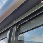 Zbliżenie na górną prowadnicę systemu Sotro, zamontowaną bezpośrednio do belki pergoli, w której osadzony jest przesuwny panel szklany w eleganckiej, ciemnoszarej ramie aluminiowej.
