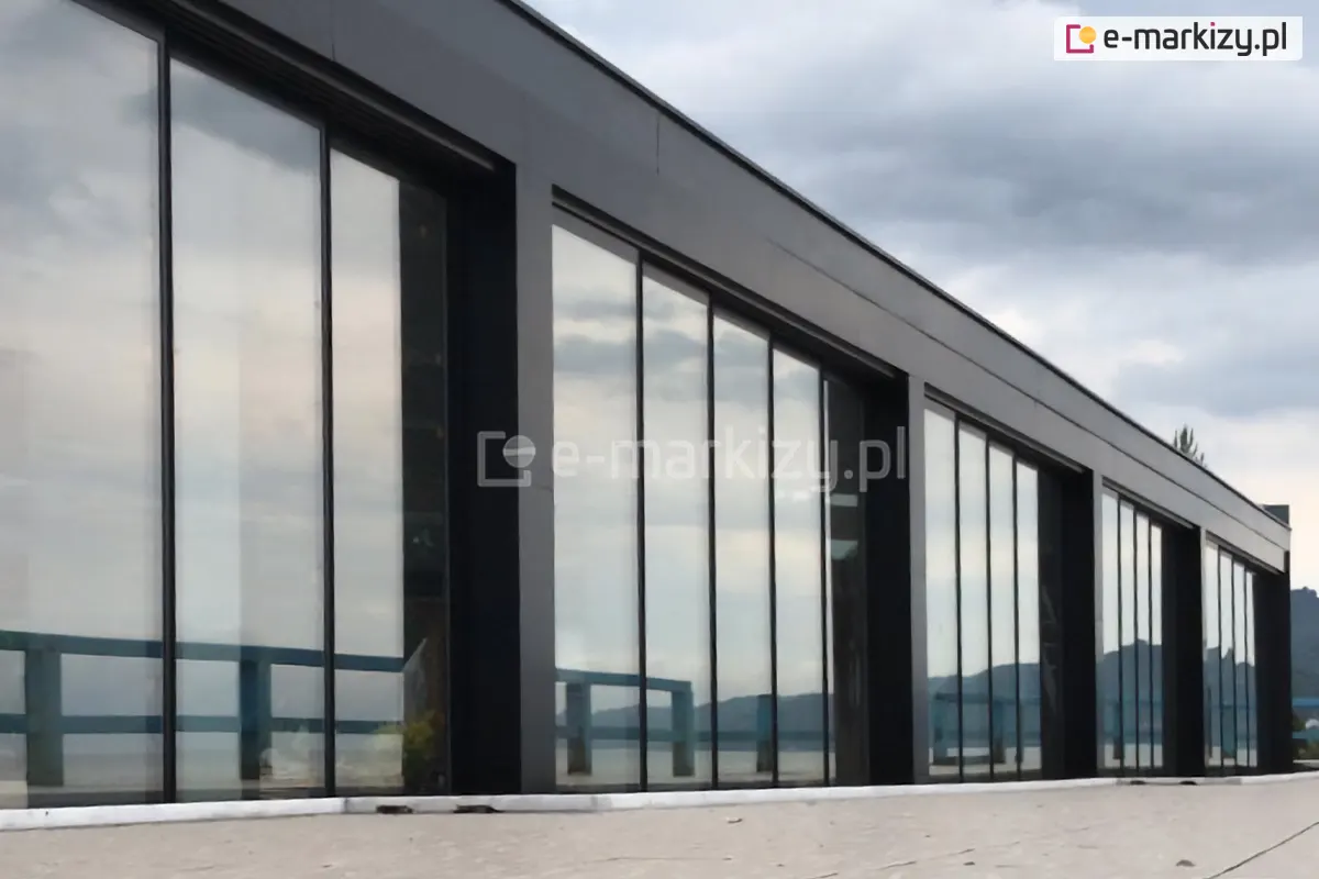 Nowoczesny budynek osłonięty na całej długości bezramowymi szklanymi ścianami przesuwnymi Miros, wykonanymi z aluminium wysokiej jakości, idealny przykład estetycznej i funkcjonalnej zabudowy tarasu lub elewacji.