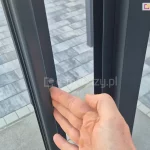 Zbliżenie na dłoń demonstrującą łatwość przesuwania lekkiego profilu systemu szklanych drzwi przesuwnych Sotro na tle szarego chodnika z kostki brukowej.