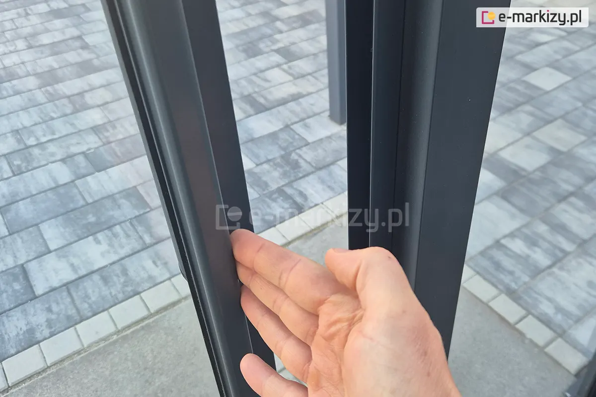 Zbliżenie na dłoń demonstrującą łatwość przesuwania lekkiego profilu systemu szklanych drzwi przesuwnych Sotro na tle szarego chodnika z kostki brukowej.