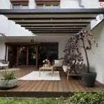 Nowoczesna antracytowa pergola tarasowa model Aura z trzema punktami LED na co drugiej belce, zamontowana na drewnianym tarasie z przytulną strefą wypoczynku i roślinnością w dużych donicach