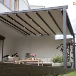 Nowoczesna pergola tarasowa Aura z rozwiniętą tkaniną chroniącą przed słońcem, pod którą ustawione są wygodna kanapa wypoczynkowa, fotel i drewniany stół w otoczeniu ciepłolubnych roślin