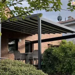 Aluminiowa pergola ogrodowa Square z tkaniną przeciwsłoneczną i punktowym oświetleniem LED na wieczór, montowana przy ścianie domu z cegły, zapewniająca komfortowe miejsce do siedzenia na tarasie z widokiem na ogród